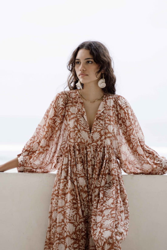 Daughters of India Kyra Midi Dress in Caramel — upper body leaning on veranda wall, V-neckline, billowed sleeves