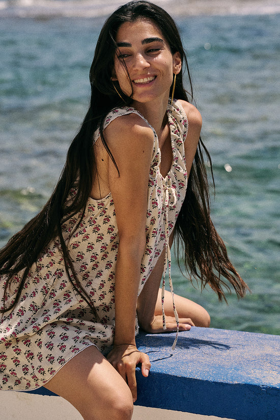 Sundress Mini Peony — seated by turquoise sea showing back tie detail, hand block print peony floral cotton mini dress, seaside lifestyle | Daughters of India