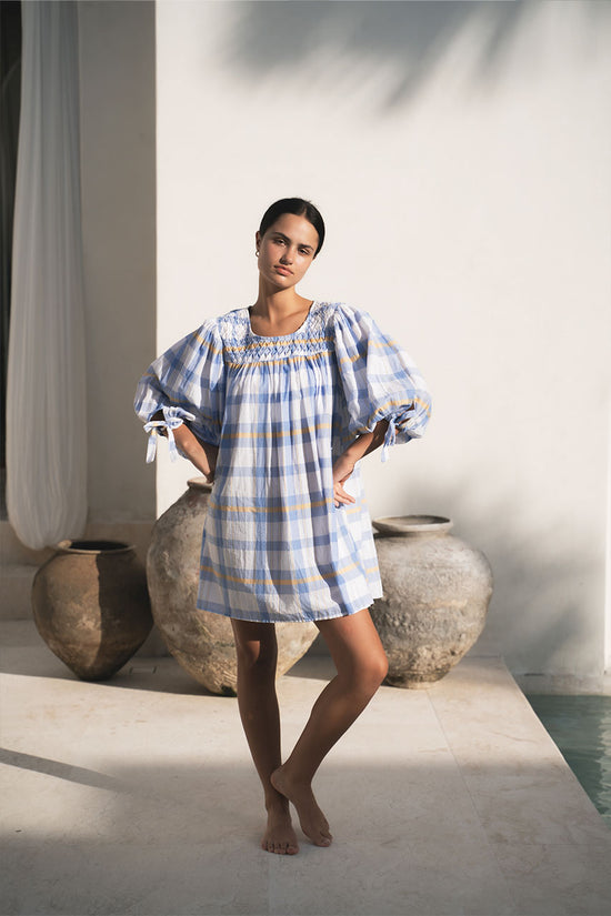 Freya Dress Periwinkle Blue Plaid — full length by terracotta pots and white walls, hand-smocked neckline, billowy sleeves, handloom woven periwinkle blue plaid mini, artisan boho dress | Daughters of India